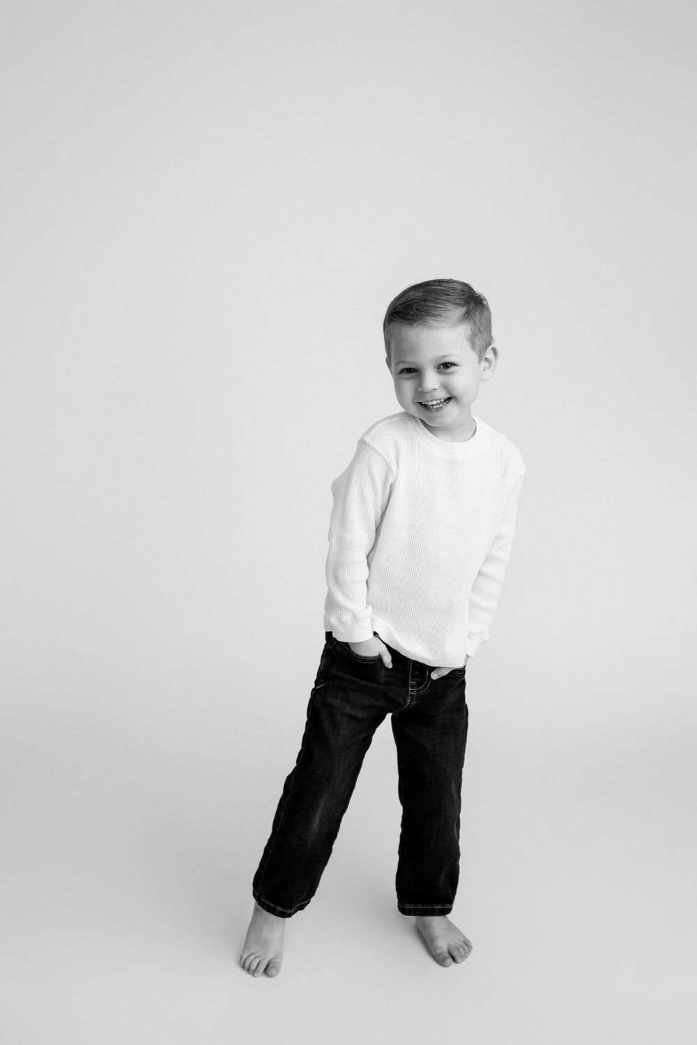 Black and white photo of a smiling young boy in white shirt and dark jeans, standing barefoot against white background.