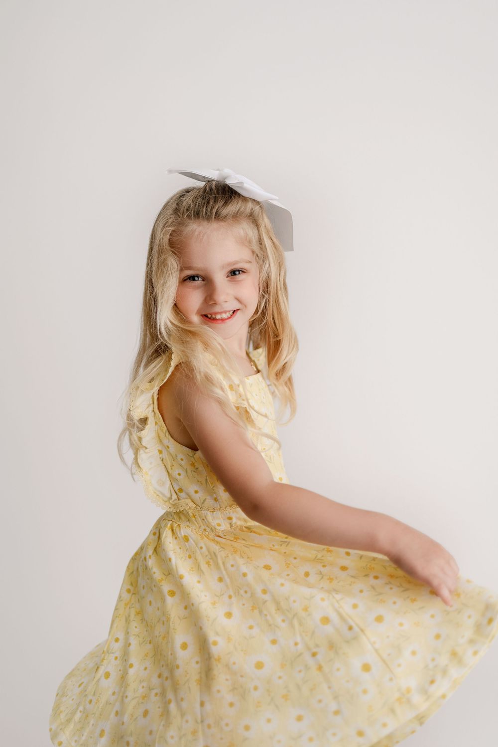 Young girl twirling in a yellow floral dress with a white bow in her blonde hair.