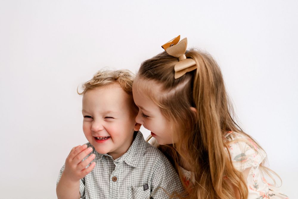Two happy children, a laughing toddler boy and smiling older girl with a bow, sharing a joyful moment together.
