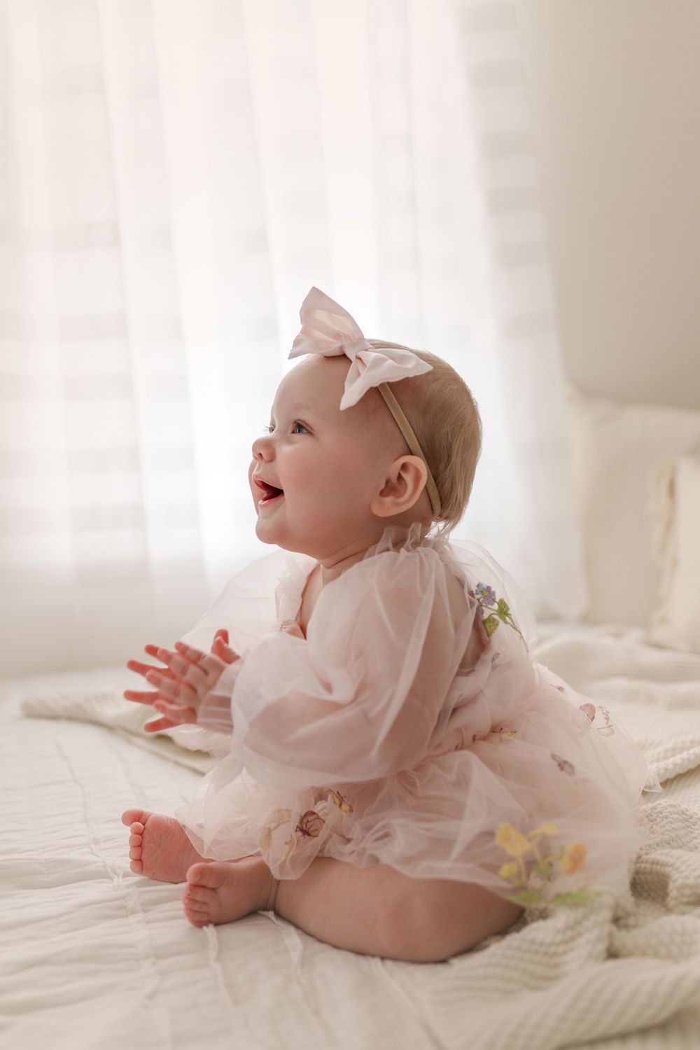A smiling baby girl in a pink tulle dress and bow headband sits on a white bed in soft natural light.