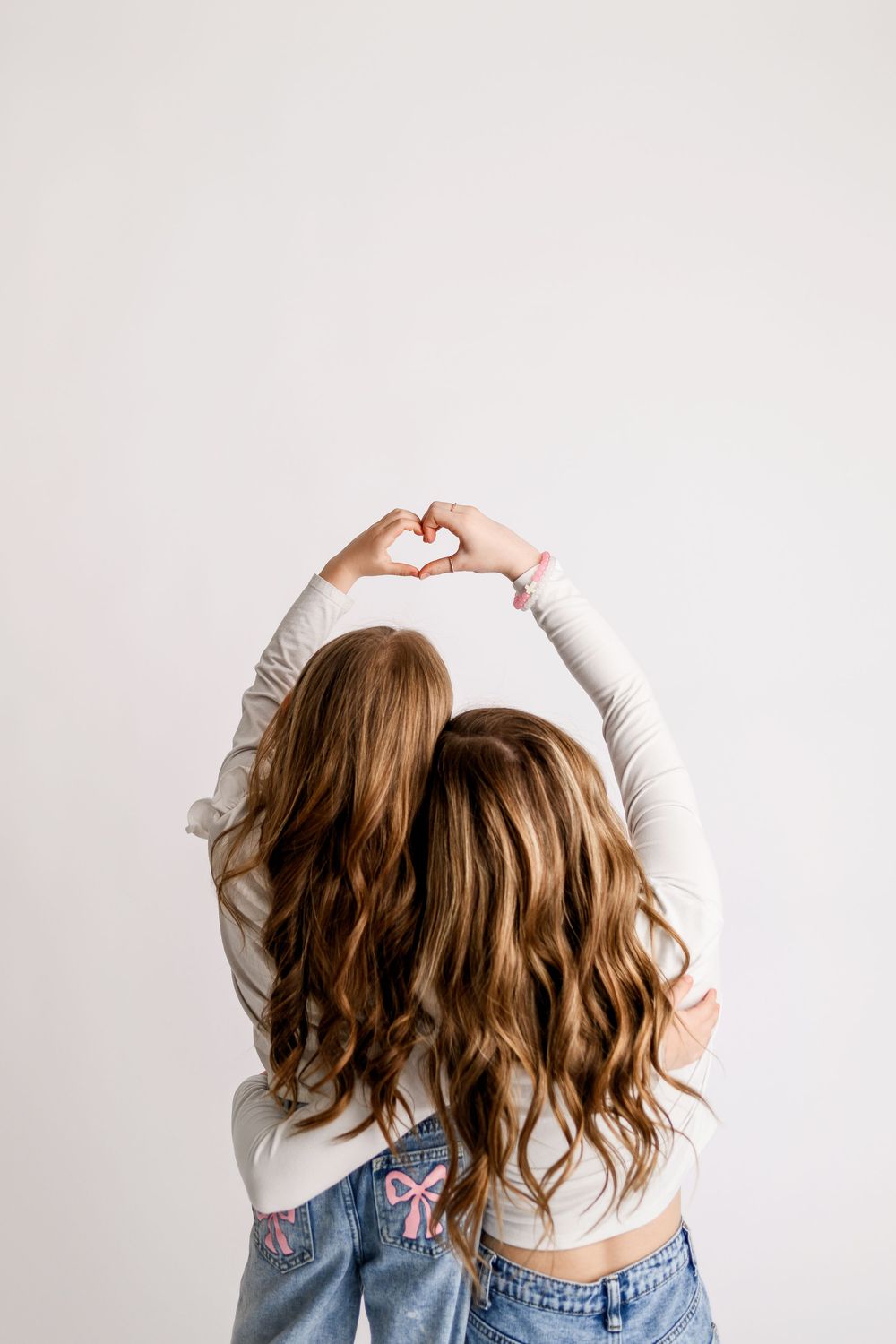 Two girls with long brown hair make a heart shape with their hands while wearing matching outfits from behind.