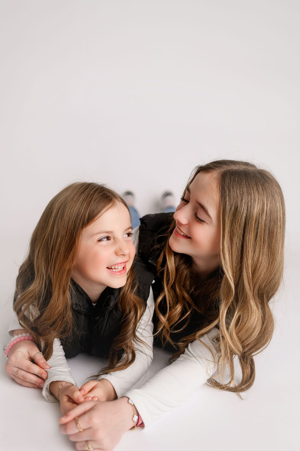 Two smiling girls with long wavy hair lying on white floor, looking at each other in a studio portrait.