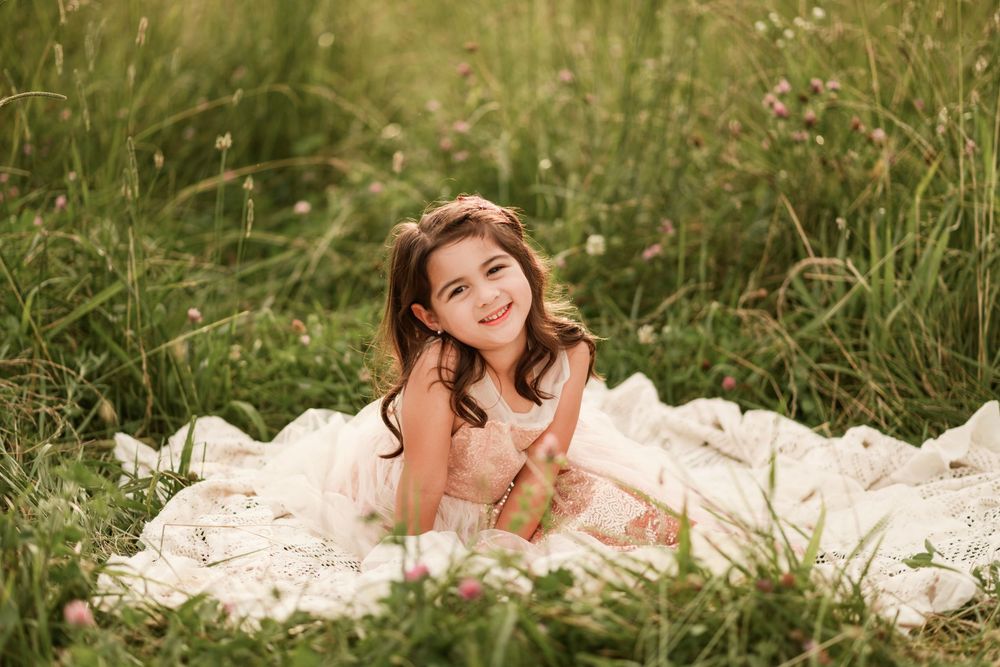 A smiling young girl in a pink dress sits on a white blanket surrounded by lush green grass and wildflowers.