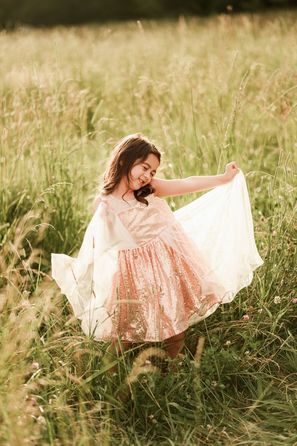 Young girl in pink sequin dress twirling in a sunlit meadow, holding white fabric with a joyful smile.