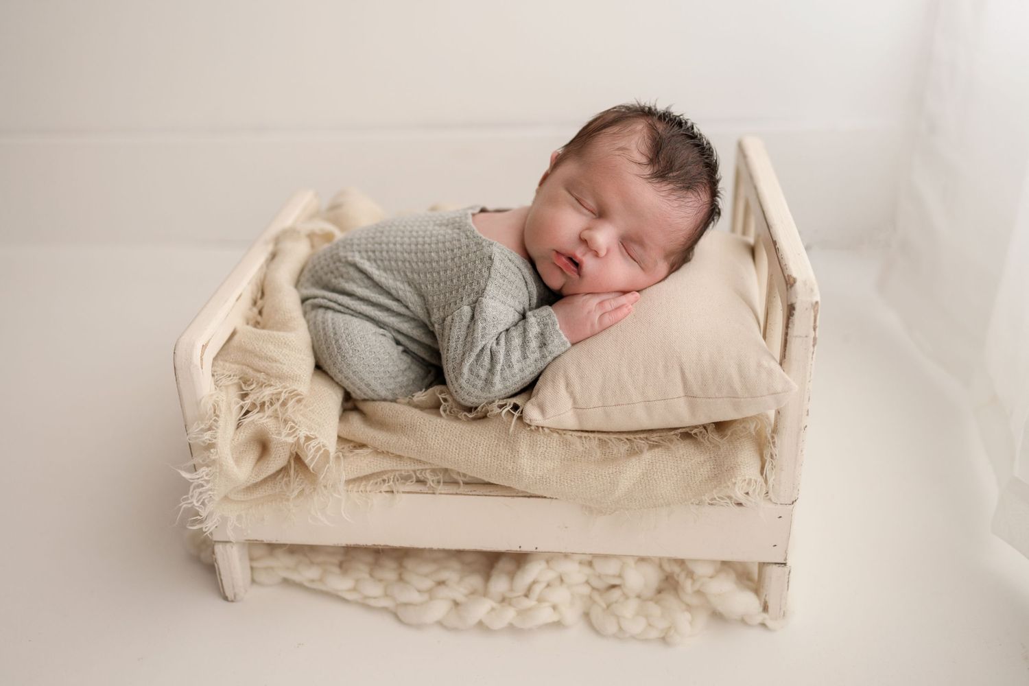 Sleeping newborn baby in gray knit outfit on cream pillow in white wooden bed prop.