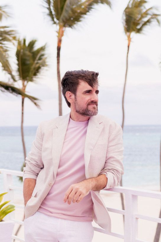 Person in light pink blazer and shirt sitting in tropical beachside setting with palm trees.