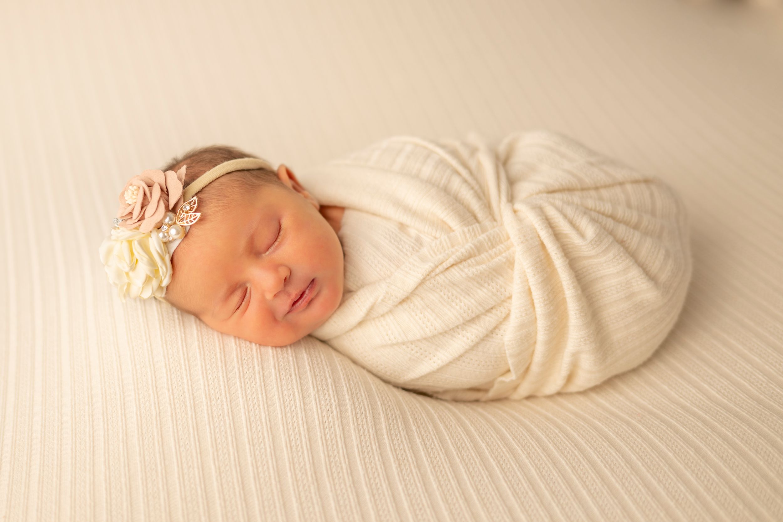 A sleeping newborn wrapped in soft white fabric with a delicate floral headband rests peacefully on textured white bedding.