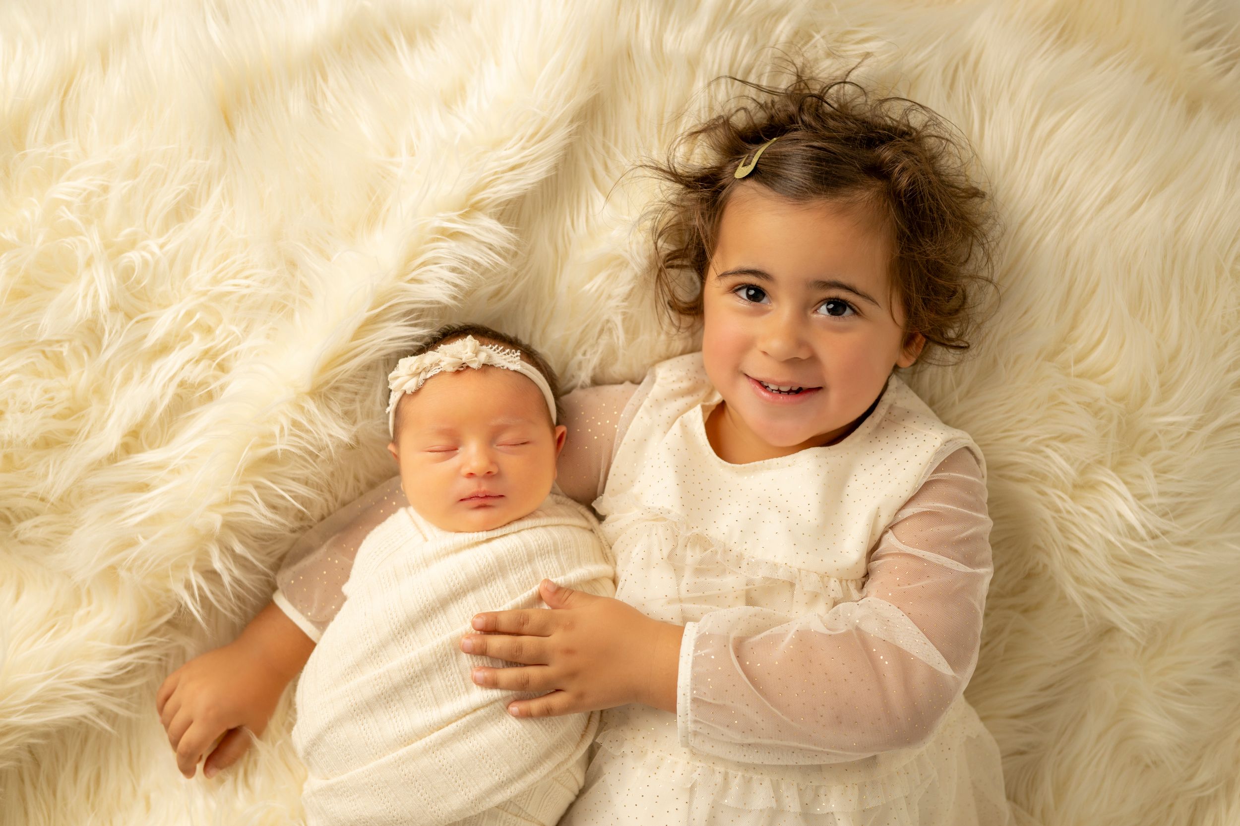 Baby wearing pink sweater lies on soft white fur blanket while smiling and playing.