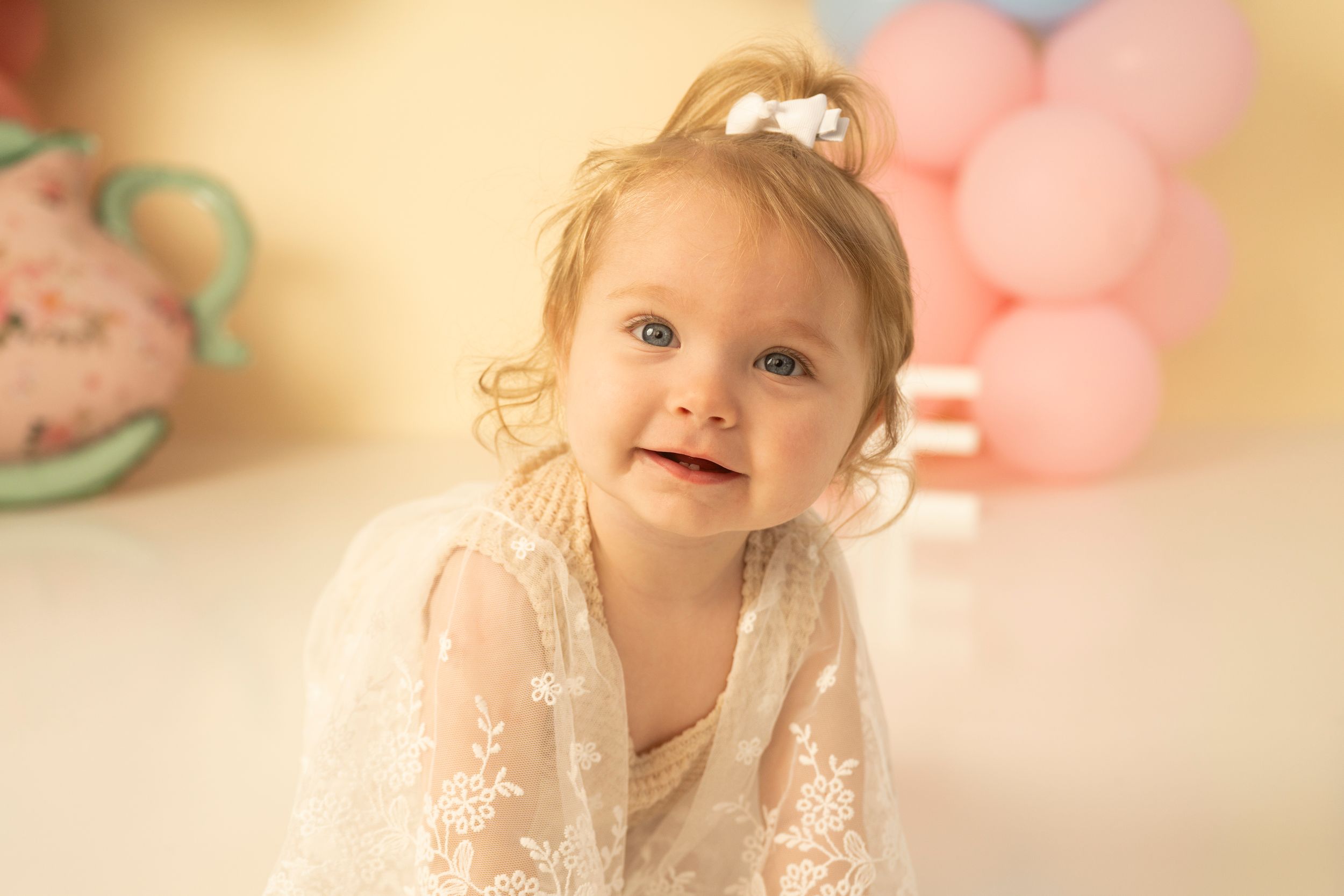 A baby in a delicate lace dress smiles while sitting against a soft pink and cream colored background.