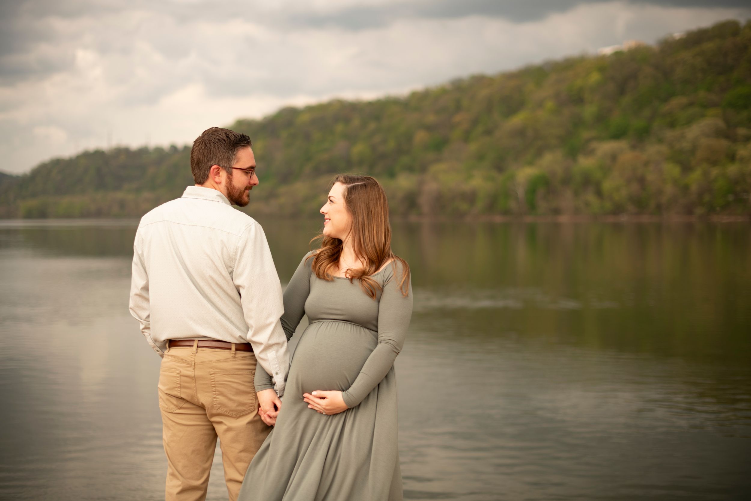 A couple in casual attire stands at the edge of a serene lake surrounded by green hills on a cloudy day.