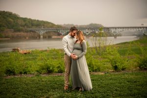 A romantic couple sits on a grassy hill overlooking a scenic river and bridge during golden hour.
