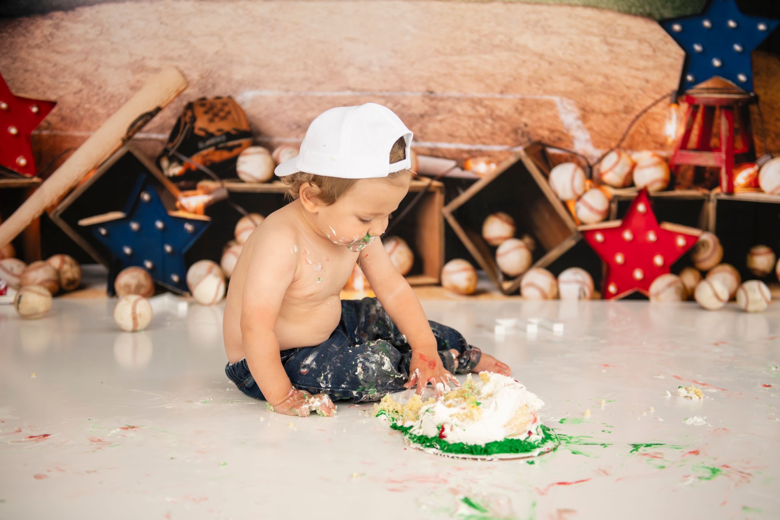 Baby in baseball cap smashing cake during first birthday photoshoot.