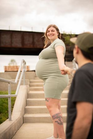 A pregnant woman in a green top poses playfully on concrete steps outdoors.