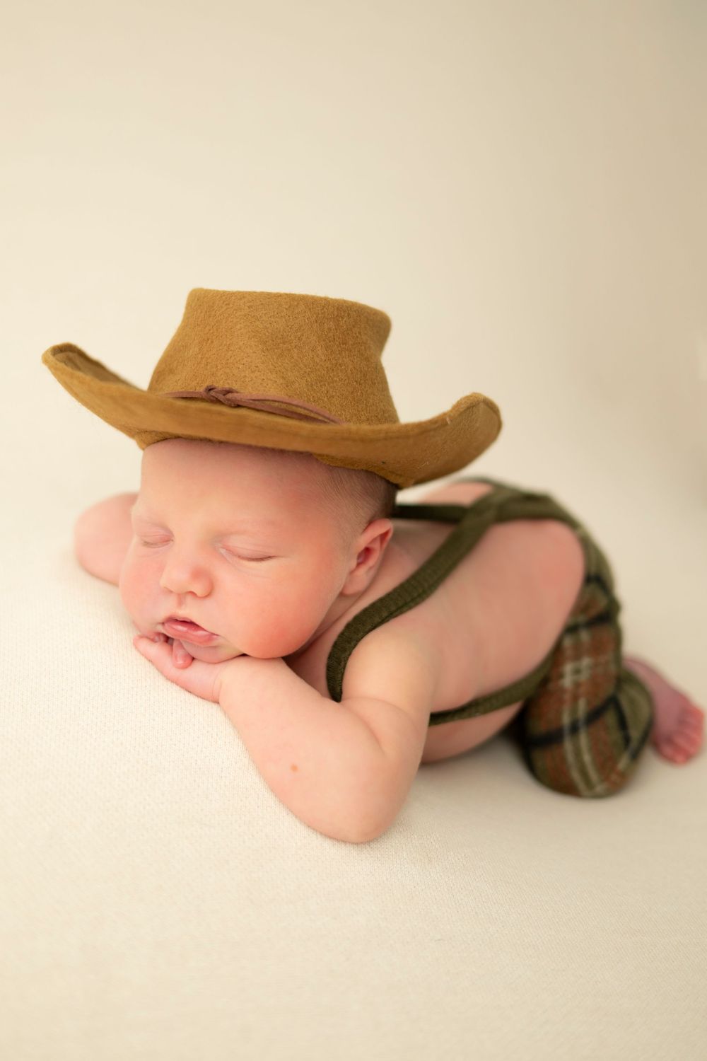 A sleeping baby wearing a brown cowboy hat and plaid shorts lies peacefully on a white background.