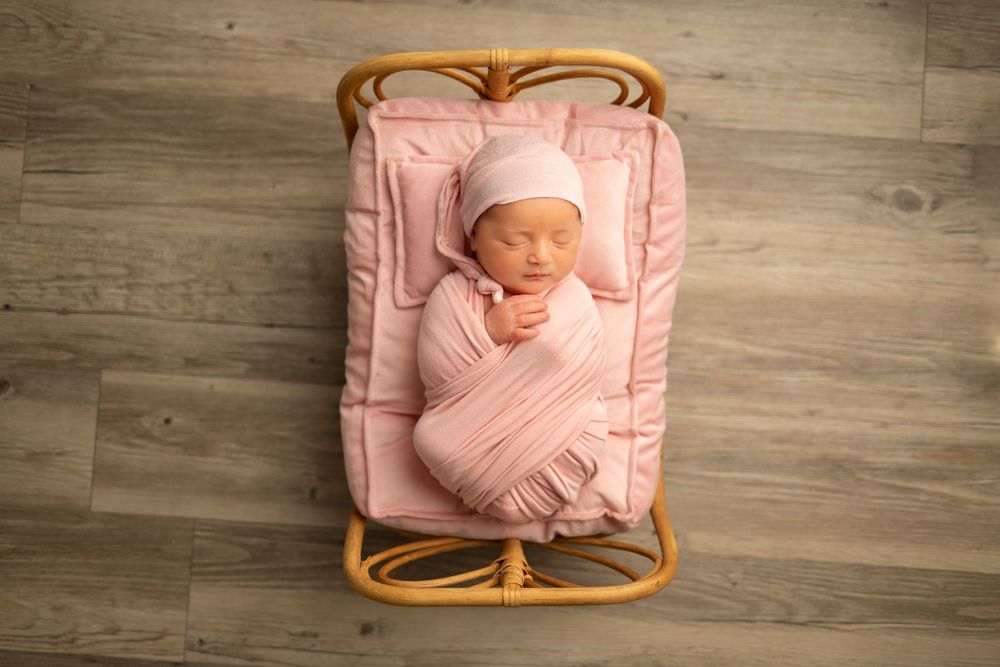 A sleeping newborn baby swaddled in pink fabric lies in a wooden basket on a gray wood floor.