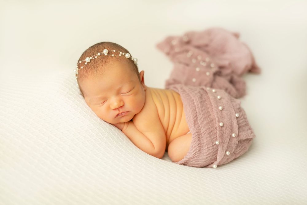 Sleeping newborn baby wrapped in soft pink swaddle blanket curled up in peaceful slumber on white backdrop.