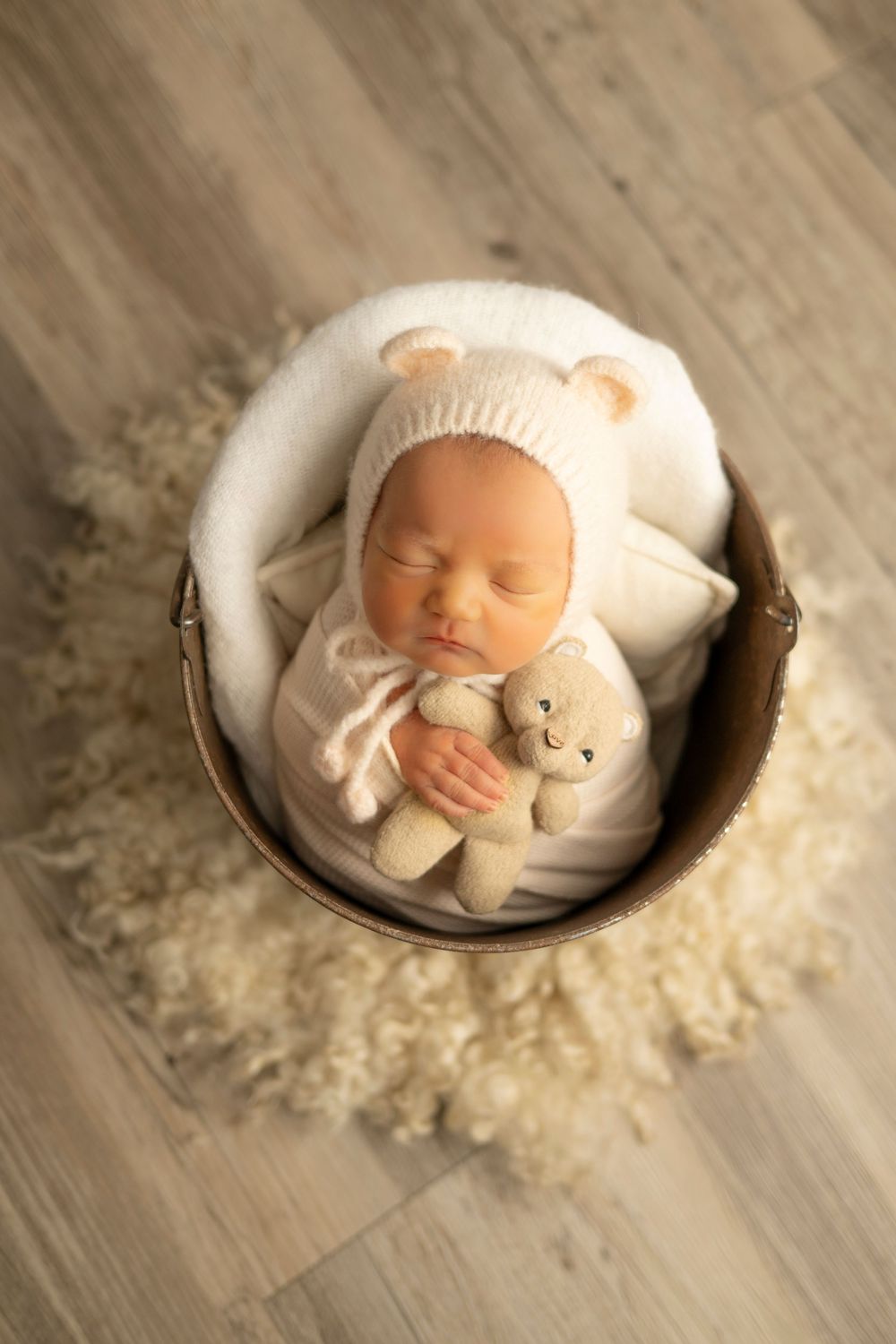 A sleeping newborn nestled in a white cushioned bowl with a plush teddy bear on a wooden floor.