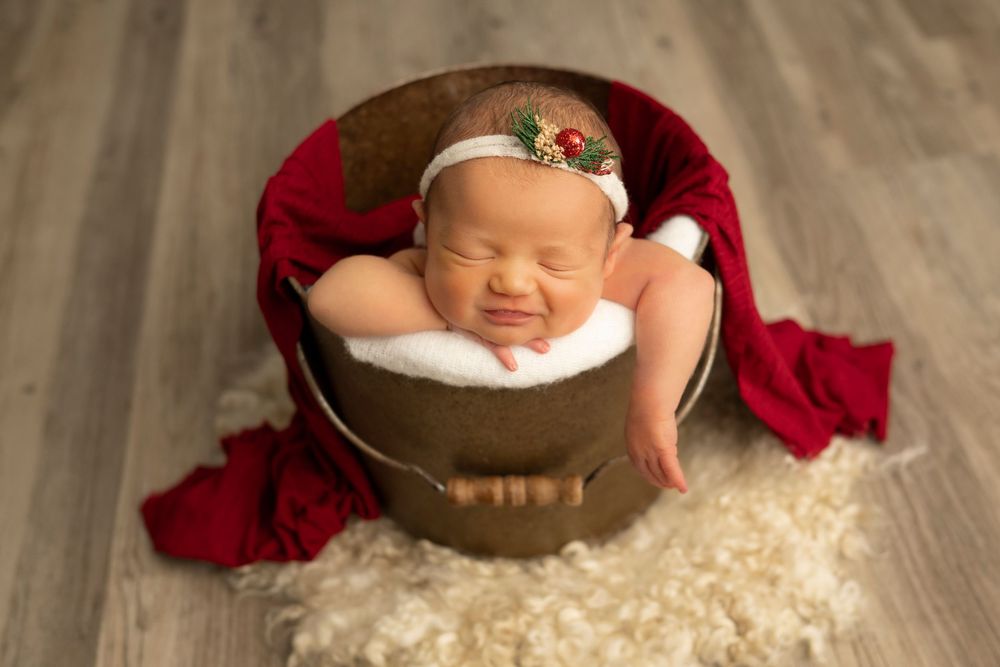 A sleeping baby nestled in a rustic wooden bowl surrounded by soft cream-colored fabric and red accents.