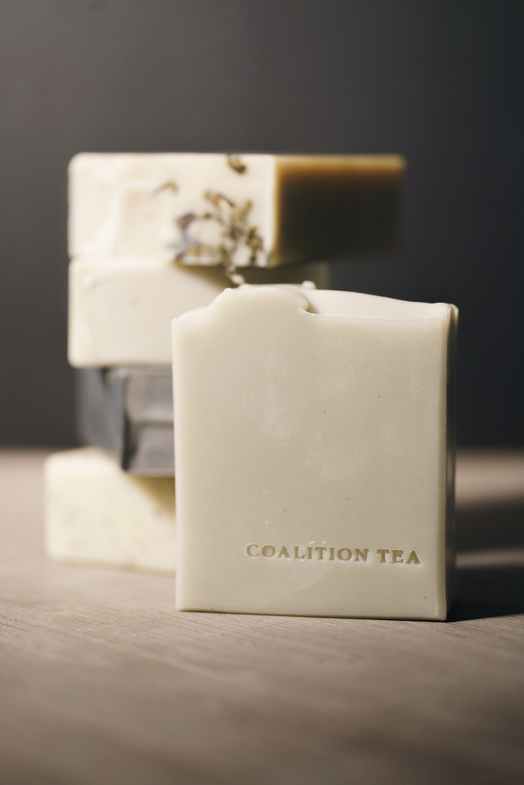 Coalition Tea branded artisan soap bars stacked on a wooden surface, showcasing natural handcrafted soaps in cream and dark tones.