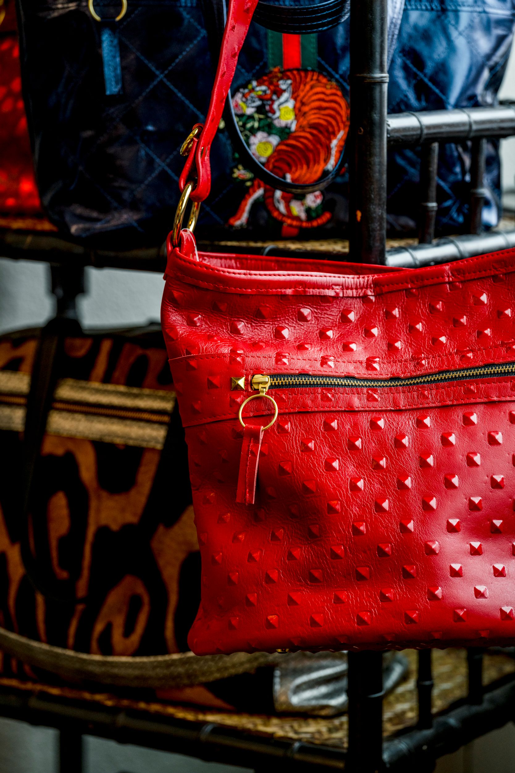 Red ostrich leather crossbody bag with gold zipper and studs displayed on a rack with other designer bags.
