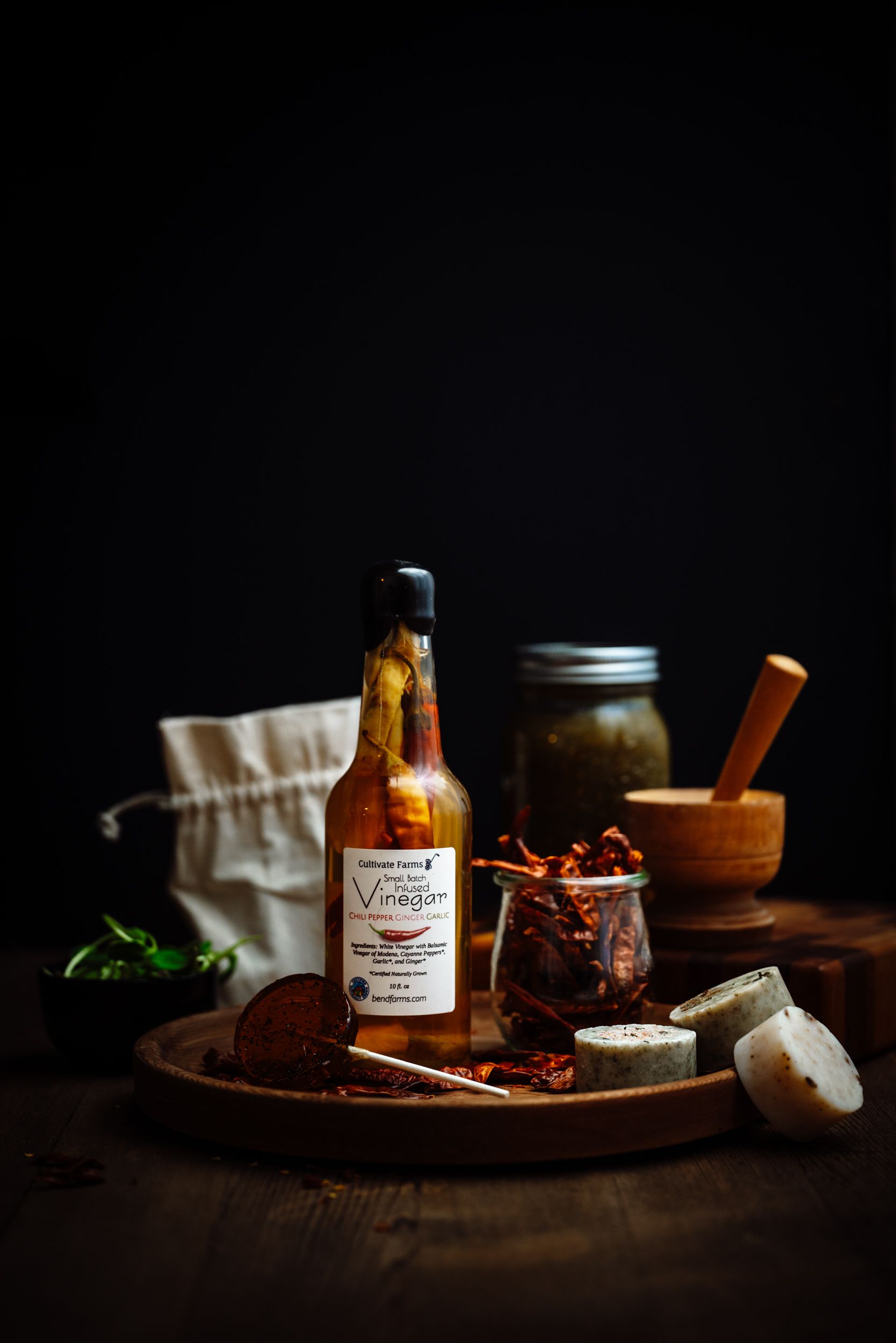Artisan herb vinegar bottle on wooden board with spices, preserved vegetables, mason jar, and mortar in moody dark setting.