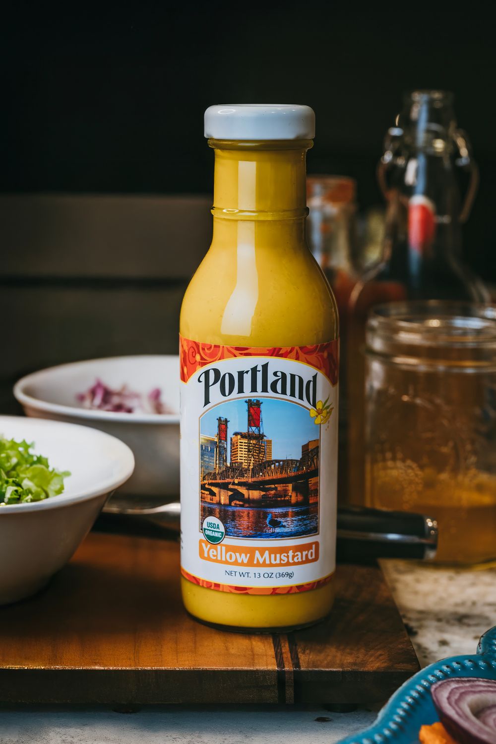 A bottle of Portland yellow mustard with a cityscape label sits on a wooden table beside bowls of food and a glass.