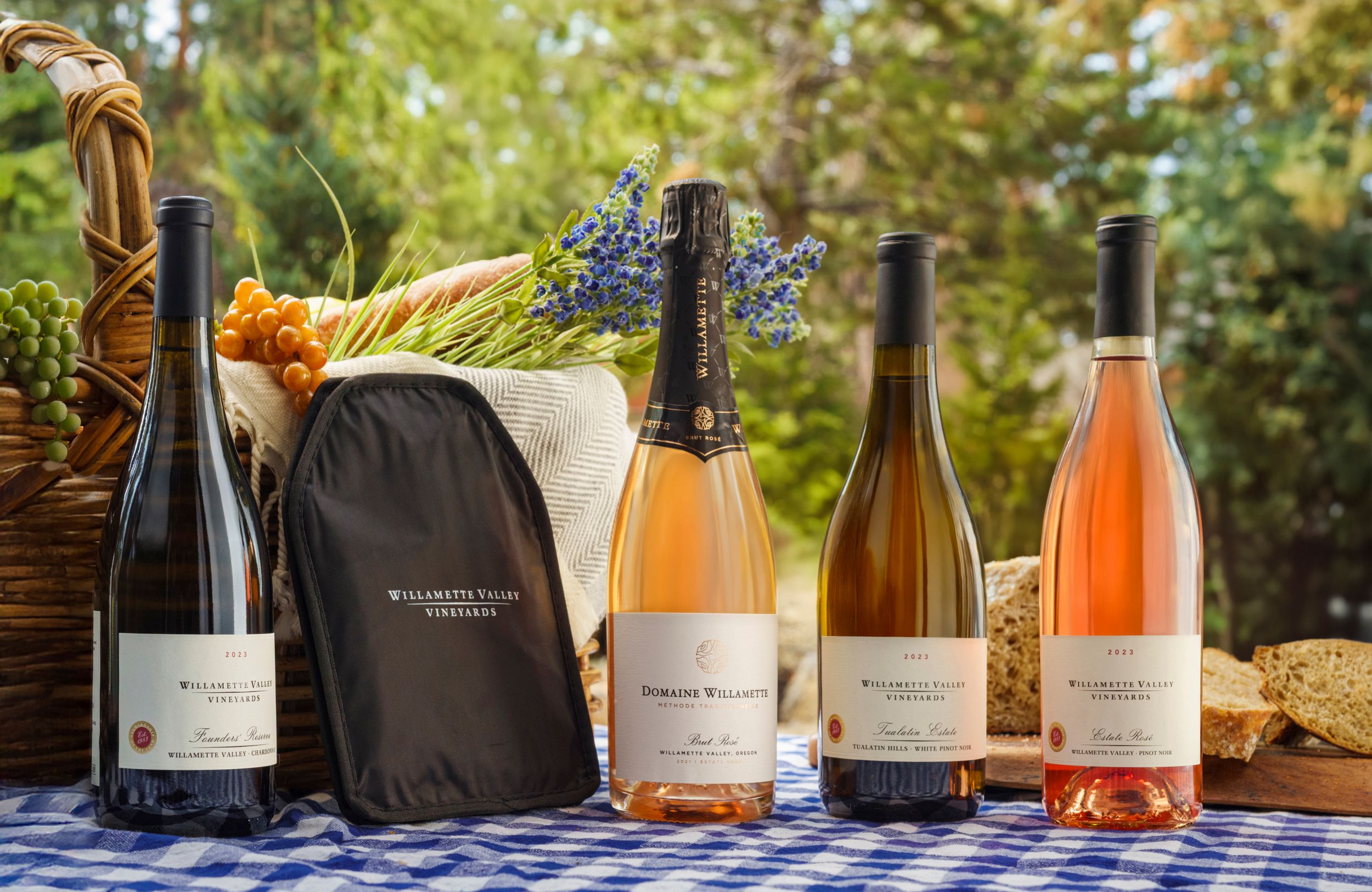 Various wine bottles including rosé and white varietals arranged outdoors with a woven basket and gray bag in natural lighting.