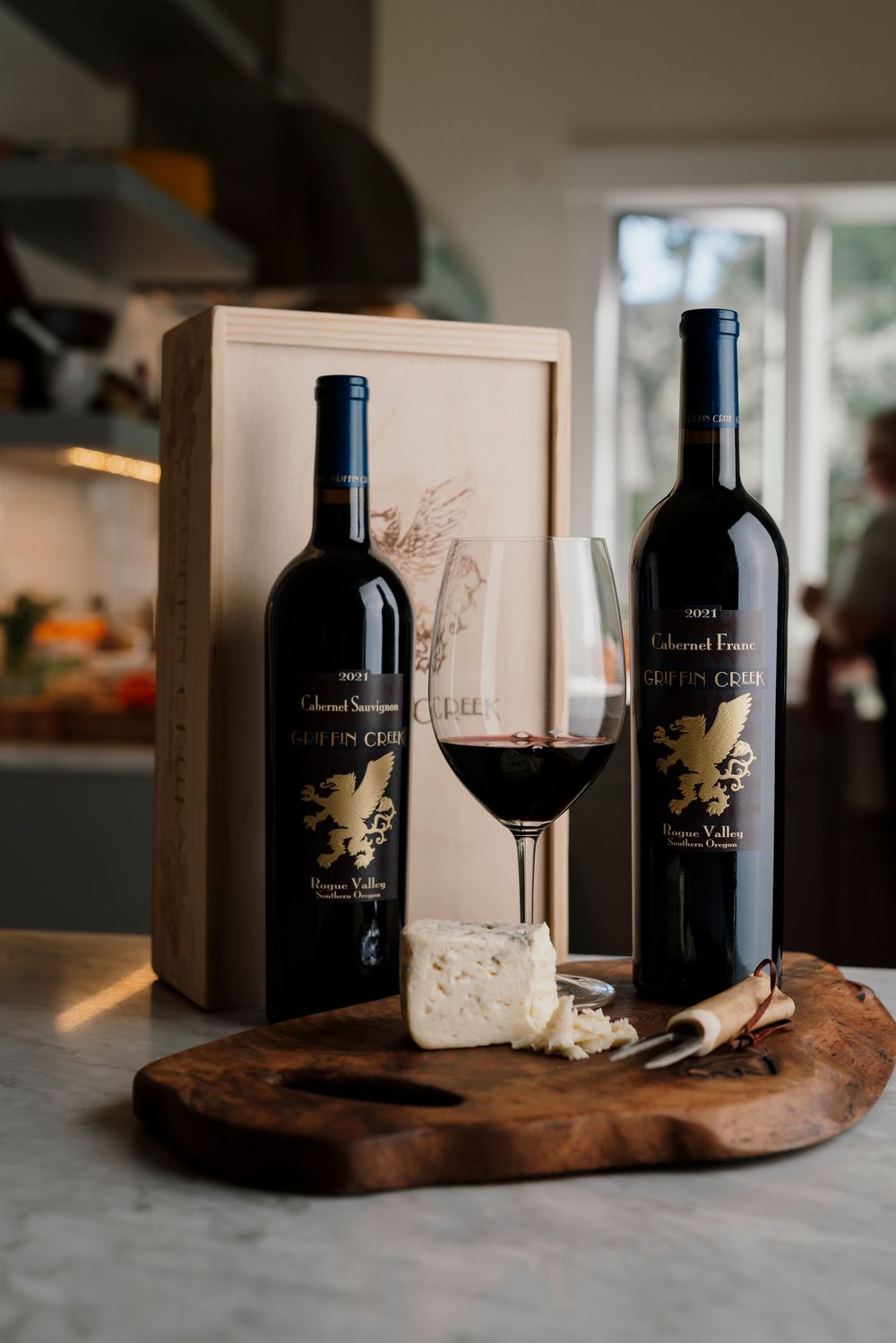 Two bottles of Griffin Creek Cabernet Sauvignon with a poured glass and cheese on a wooden board.