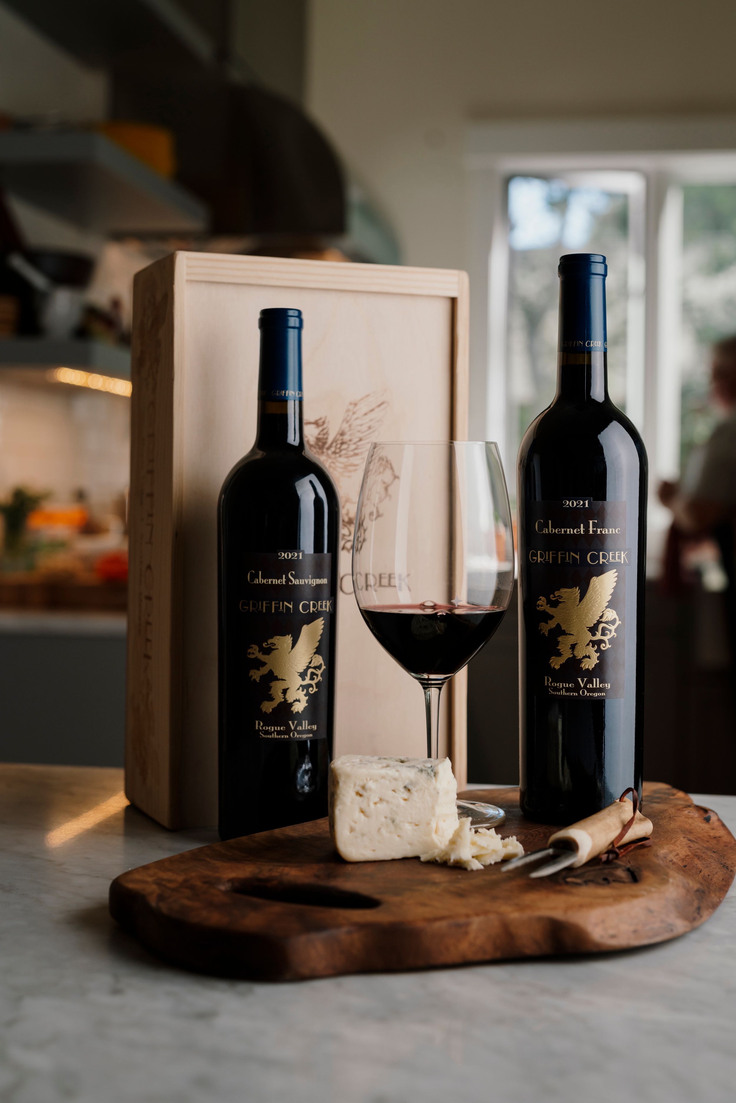 Two bottles of Griffin Creek Cabernet Sauvignon with a poured glass and cheese on a wooden board.