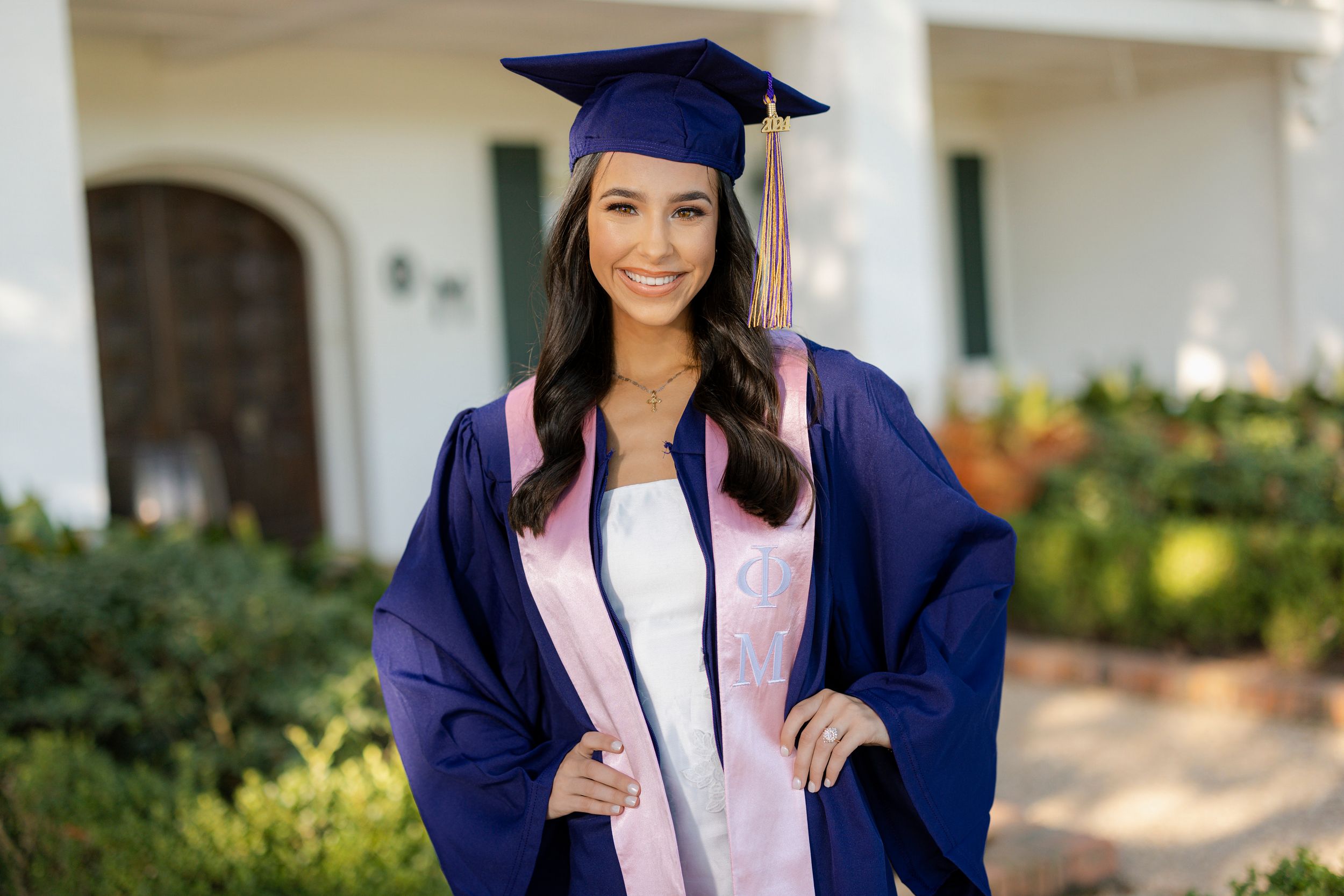 A graduate in navy blue cap and gown with pink stole poses outdoors on sunny graduation day.