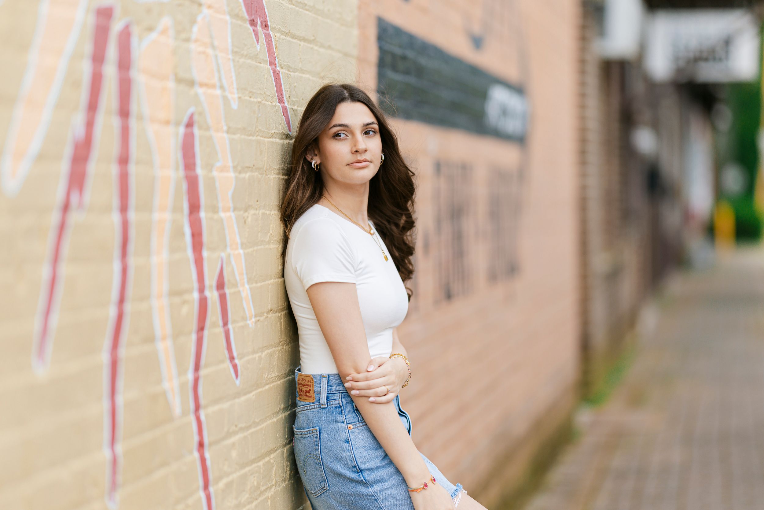 A woman in a casual white t-shirt and denim outfit poses outdoors against an urban wall.