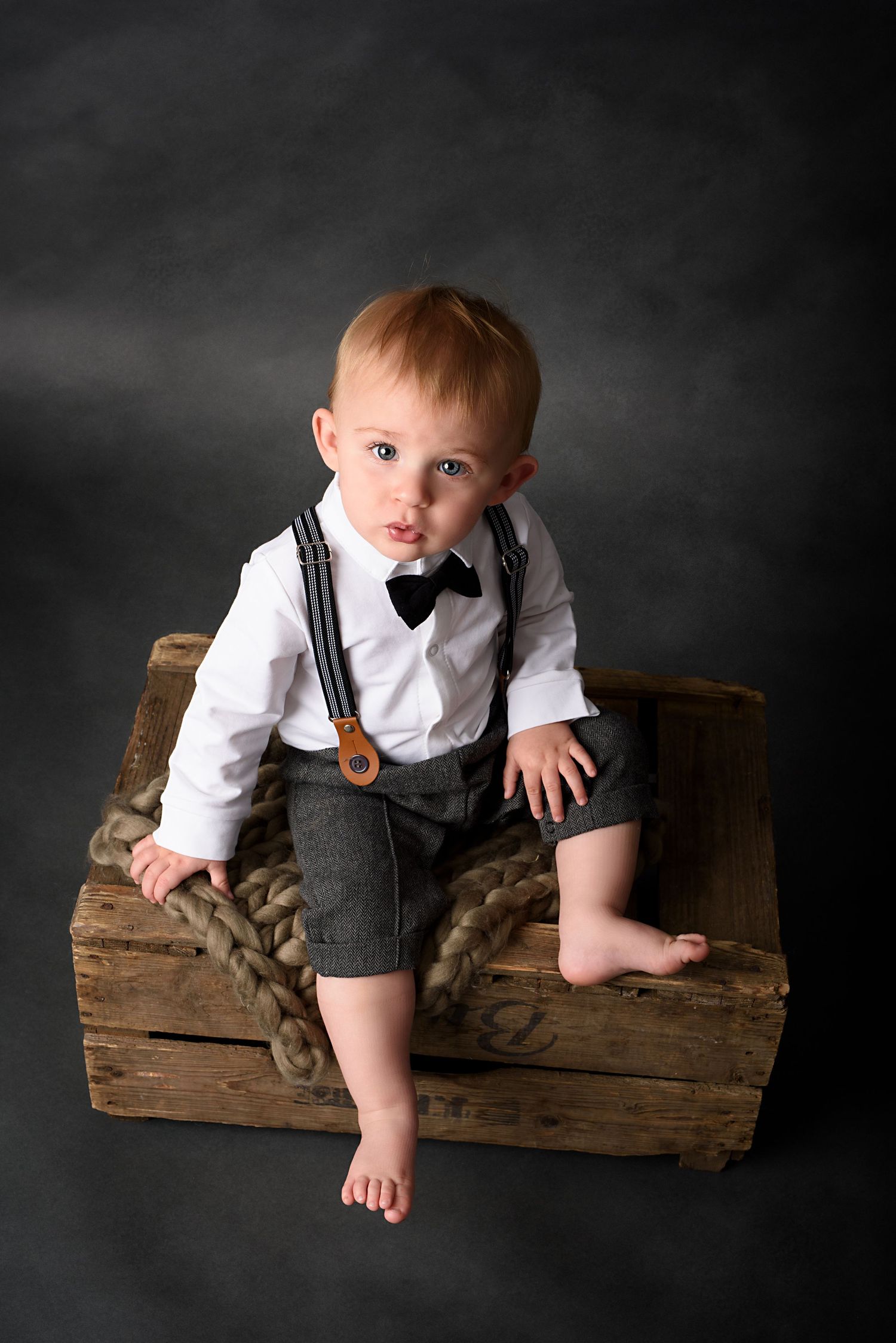 Baby in formal grey suspender outfit poses on rustic wooden crate against dark background.