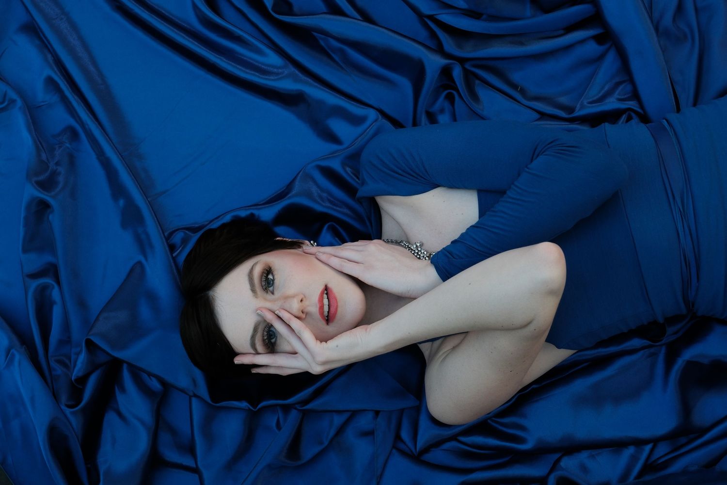 Woman with dark hair lying in blue satin sheets looking up at camera with dramatic lighting.