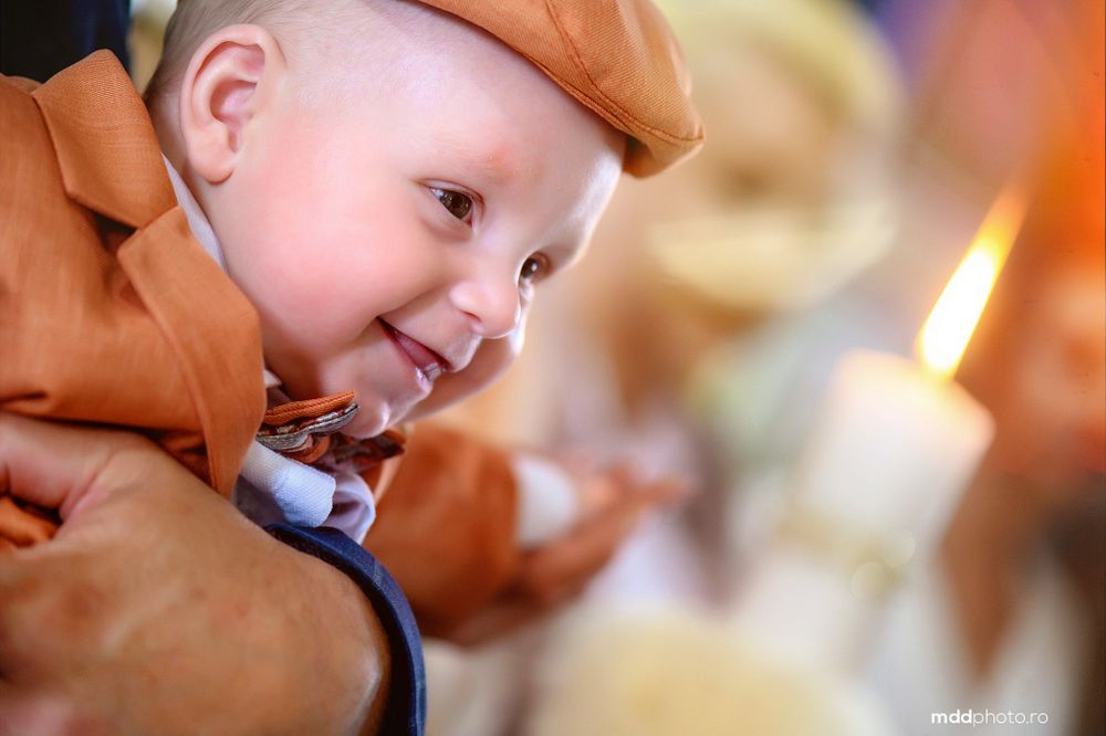 A young child in a brown coat smiles while looking at a glowing candle in soft focus.