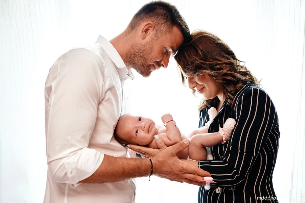 Parents in white and pinstriped clothing share a tender moment with their newborn baby against white background.