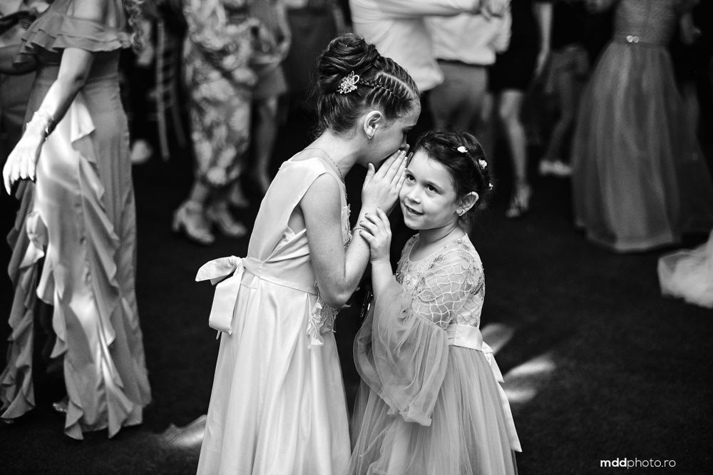 Two young bridesmaids share a sweet moment in elegant white dresses at a wedding reception in black and white.