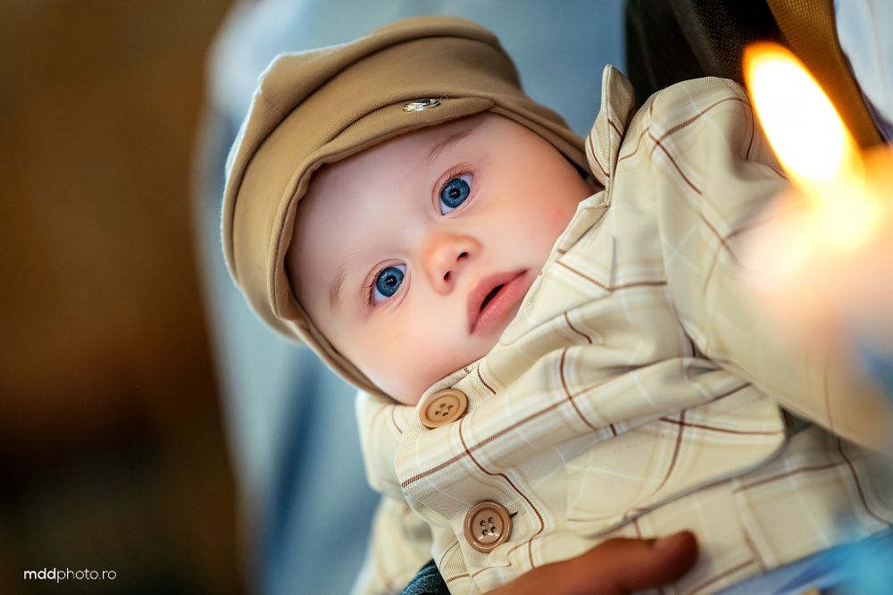 A baby wearing a beige cap and matching outfit looks up with bright blue eyes as candlelight glows nearby.