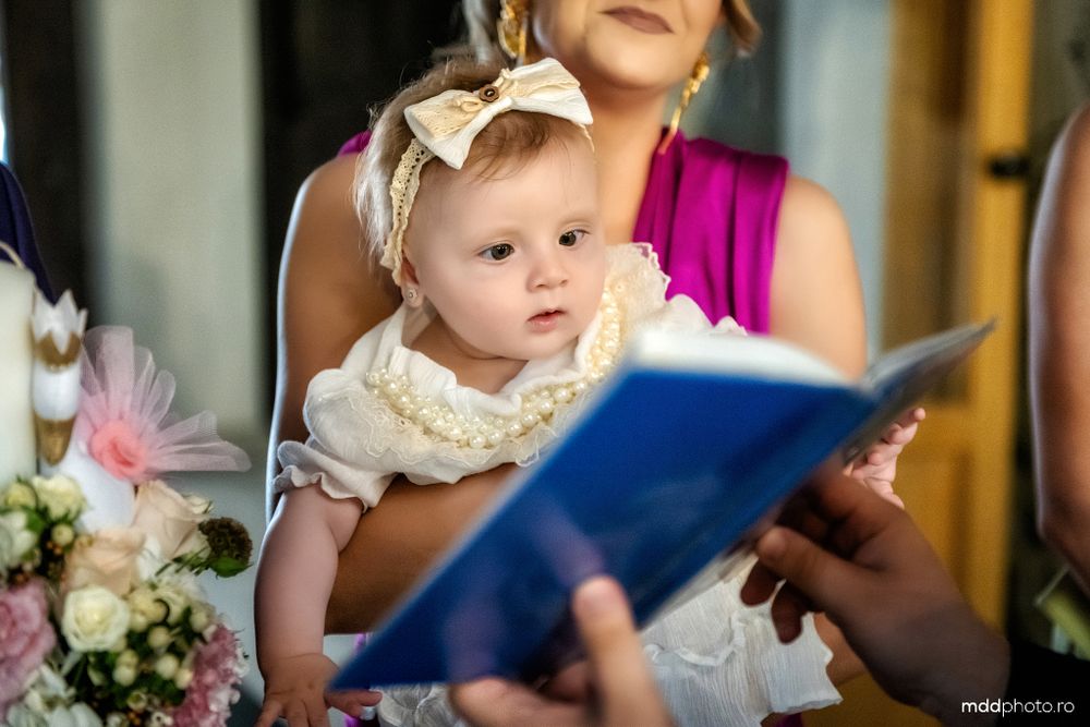 A baby in a white lace dress looks on as someone reads from a blue book during a ceremony.