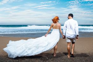 A couple walks on a black sand beach with a flowing white wedding dress against blue ocean waves and sky.