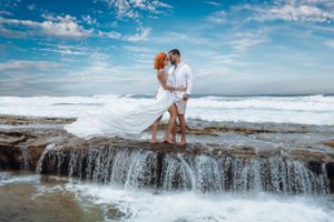 Romantic couple poses on coastal rocks with crashing waves and a flowing white dress at the beach.