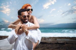 Joyful couple in white outfits sharing a playful moment by the ocean.