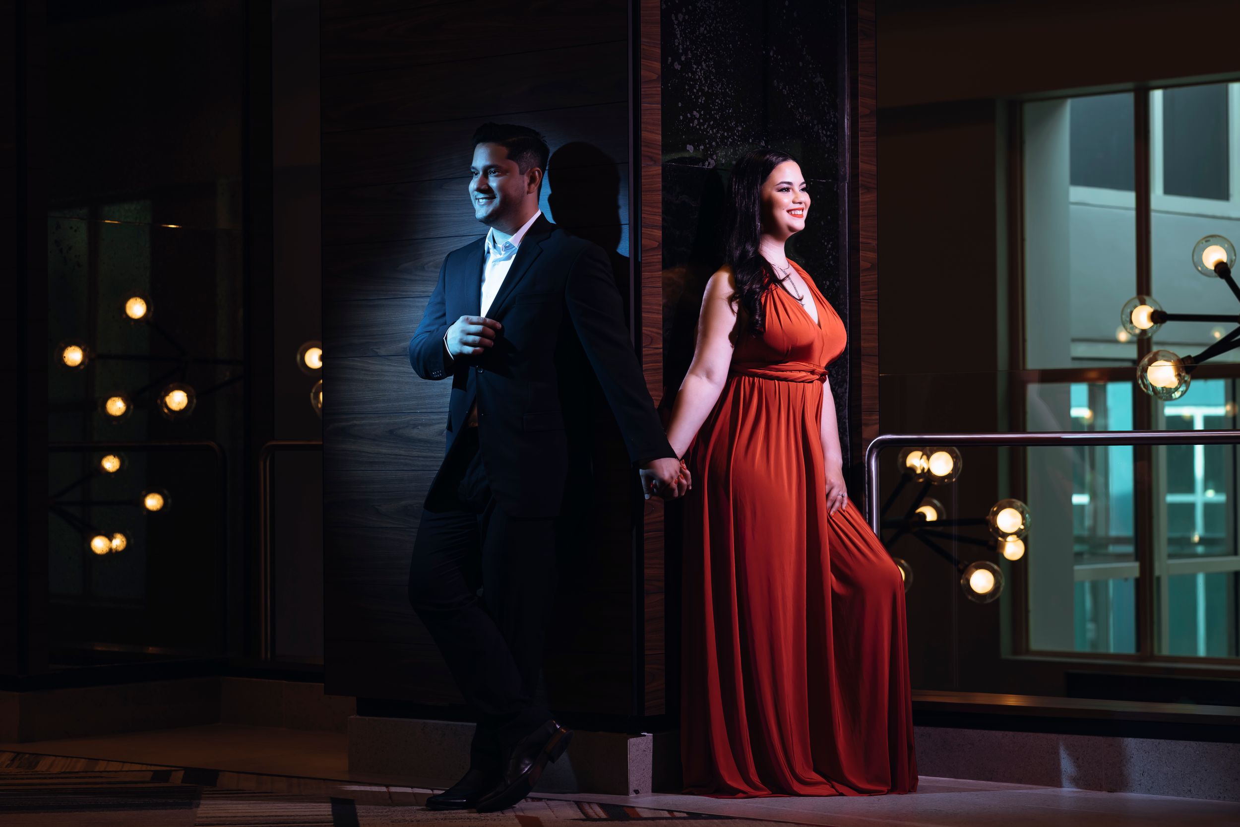 Elegant couple in formal wear pose dramatically against lit window display at night.