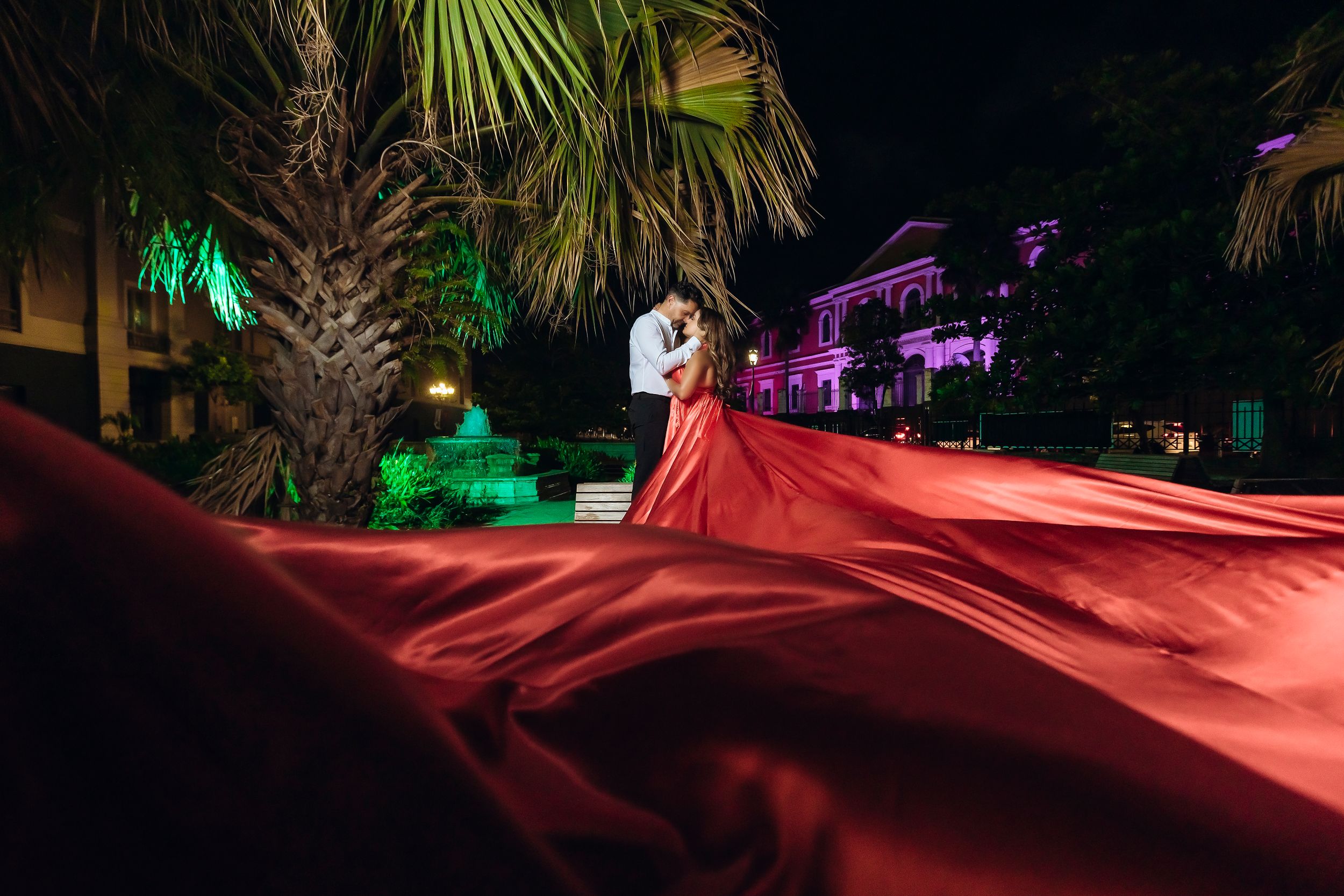 Dramatic nighttime portrait with flowing red dress fabric against palm trees lit with colored lights.