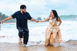 A couple plays joyfully in the ocean waves at sunset wearing navy and tan colored outfits on a sandy beach.