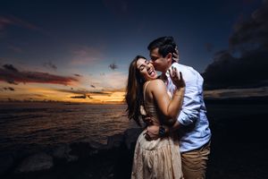 A romantic couple embraces on a beach at sunset with dramatic orange sky and dark clouds over the ocean.