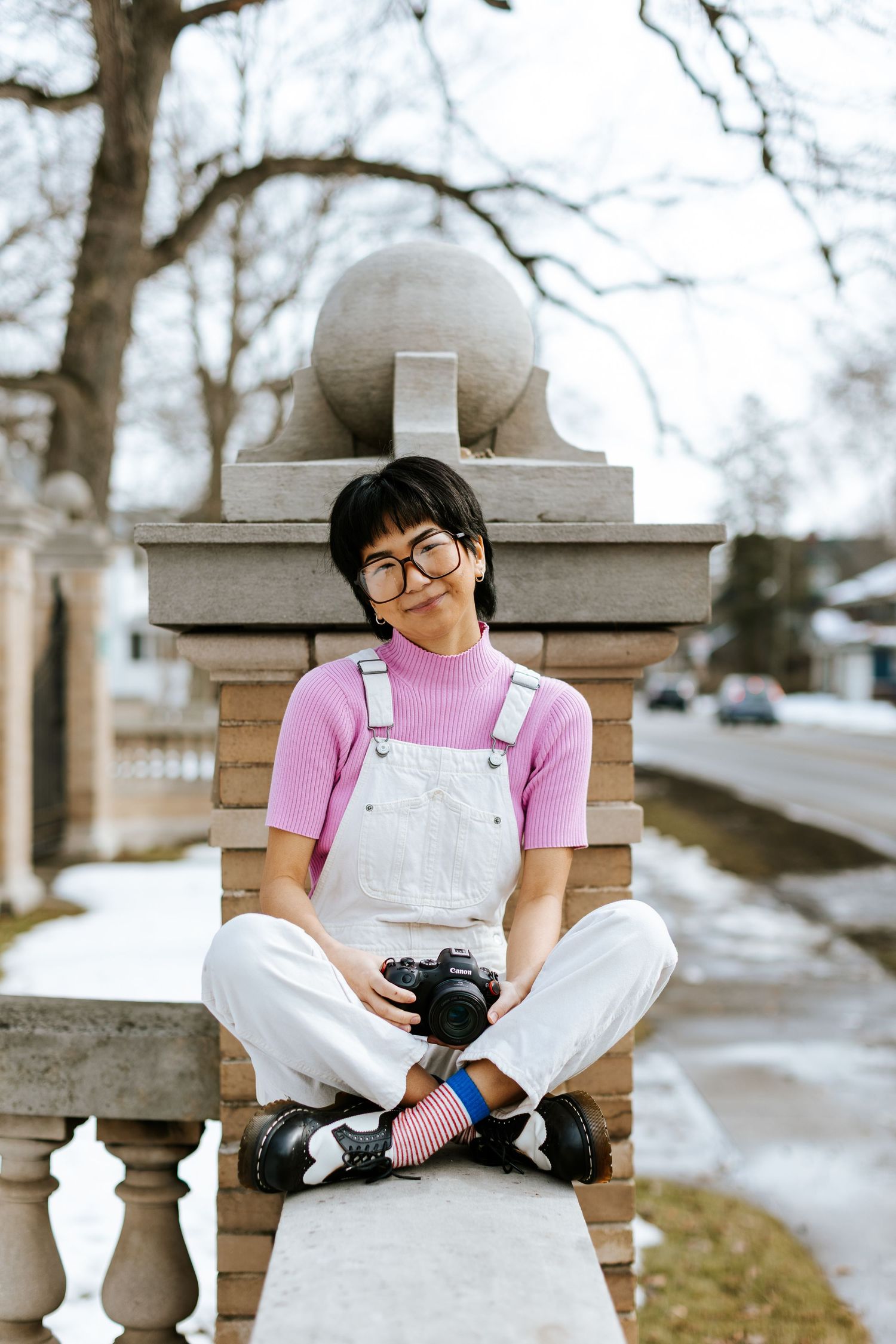 Young photographer in denim dress holding camera under willow tree branches.