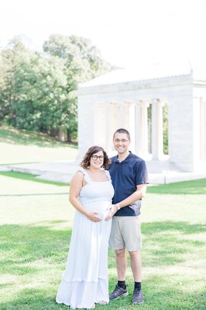 A couple poses together on a sunny day by a white columned building with green lawn and trees in the background.