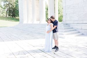 A couple embraces on the steps of a white stone monument with columns and green trees in the background.