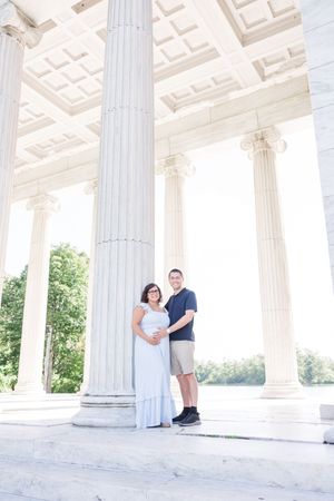 A couple stands among elegant white Grecian columns in a classical architectural setting.