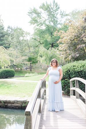 A person in a flowing white dress stands gracefully on a wooden footbridge over a tranquil pond in a garden setting.